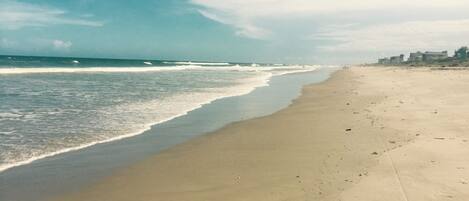A wide sandy beach with gentle waves, a clear sky, and buildings in the distance.