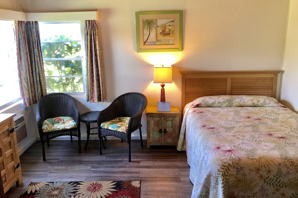 A room with a bed, two wicker chairs, a small table, a lamp, and a window with curtains.