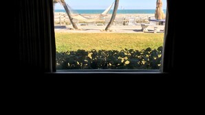 Une vue depuis une fenêtre donnant sur une plage, des palmiers et du mobilier d’extérieur.