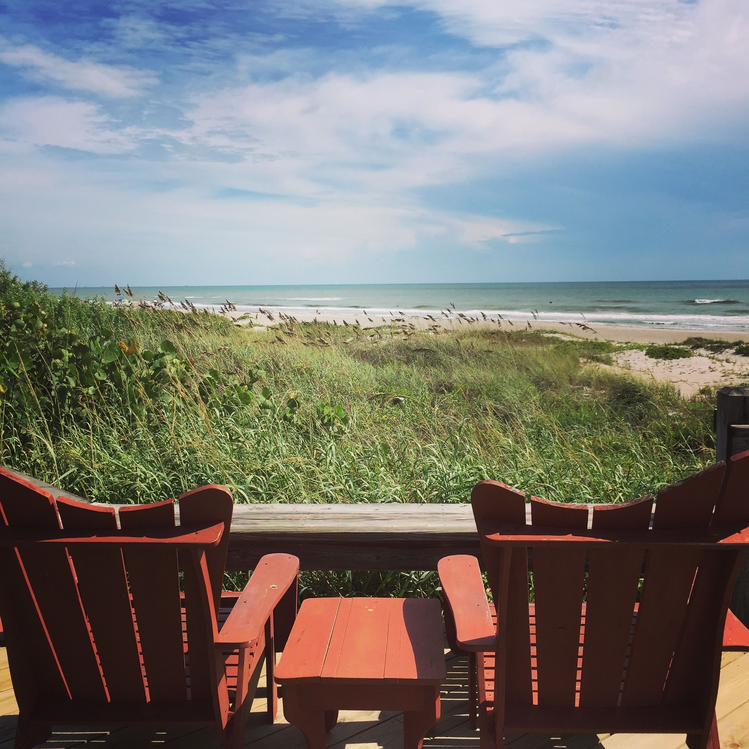 Een houten terras met rode stoelen en een tafel, met uitzicht op een strand, een grasduin en de oceaan.
