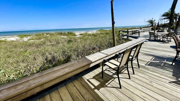 Une terrasse en bois avec un banc et des chaises, donnant sur une plage bordée de dunes et l’océan.