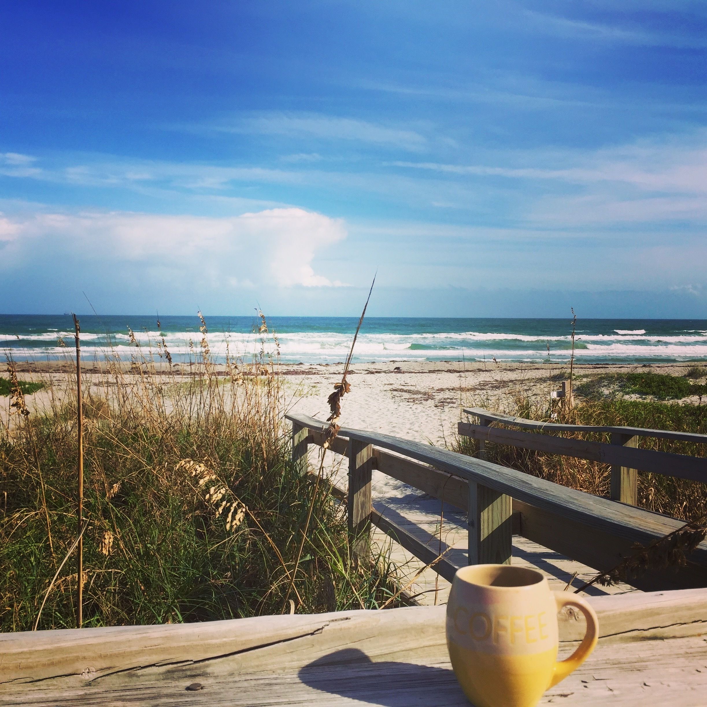 A wooden deck with a coffee cup, overlooking a beach with waves and a wooden walkway.
