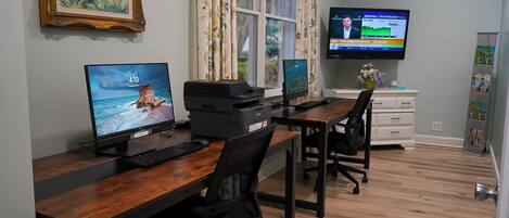 A home office with two desks, one with a desktop computer and the other with a laptop, a printer, and a TV displaying a news channel.