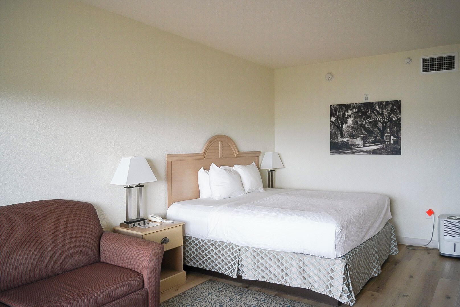 A hotel room with a bed, two bedside lamps, a nightstand, a red armchair, and a wall-mounted picture.