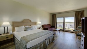 A hotel room with a large bed, two bedside tables with lamps, a sofa, a small table, and a TV. The room has a balcony with a view of the water. - Epworth By The Sea (St. Simons Island)