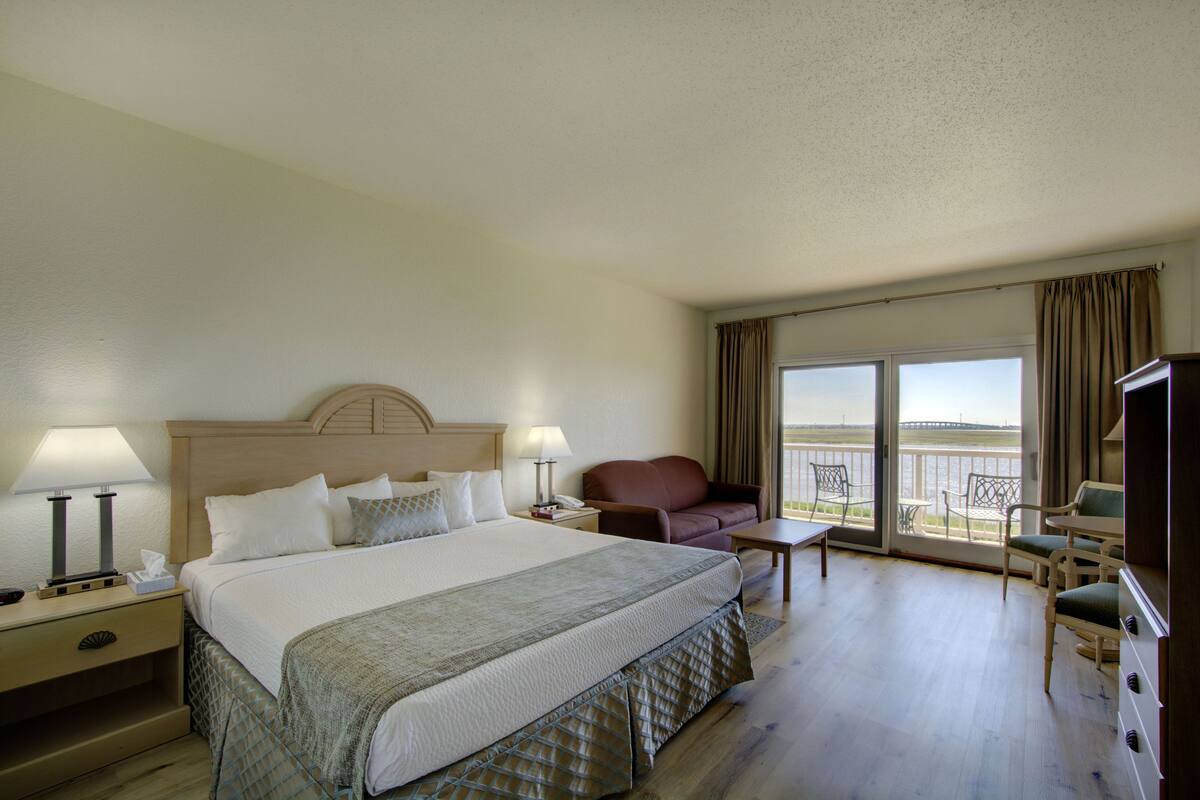 Habitación de hotel con una cama grande, dos mesitas de noche con lámparas, un sofá, una mesita pequeña y un televisor. La habitación tiene un balcón con vista al agua.