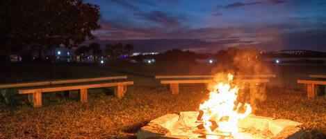 A campfire in a grassy area with picnic tables and a tree in the background.