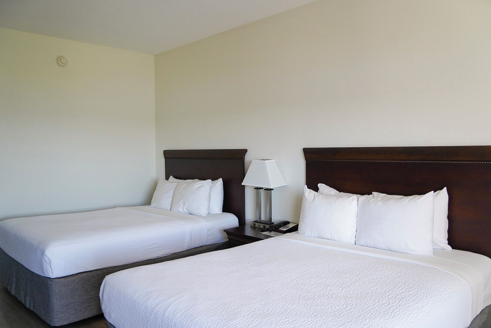 A hotel room with two single beds, a bedside table with a lamp, and a plain wall.