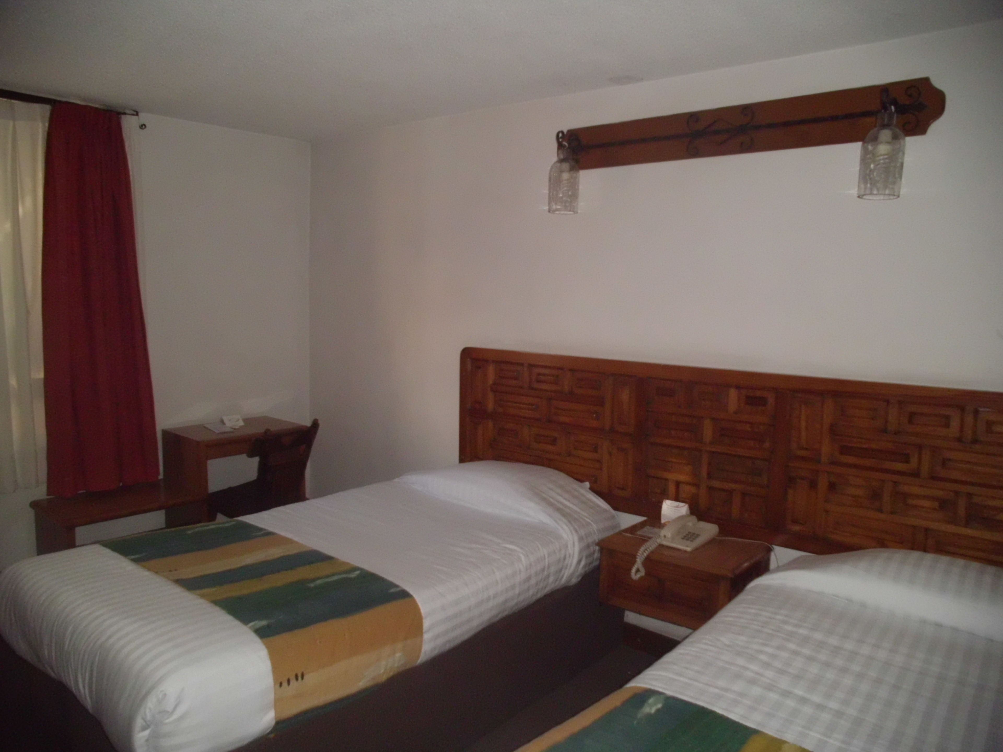 Una habitación de hotel con dos camas individuales, cabecera de madera, mesita de noche con teléfono y cortina roja.