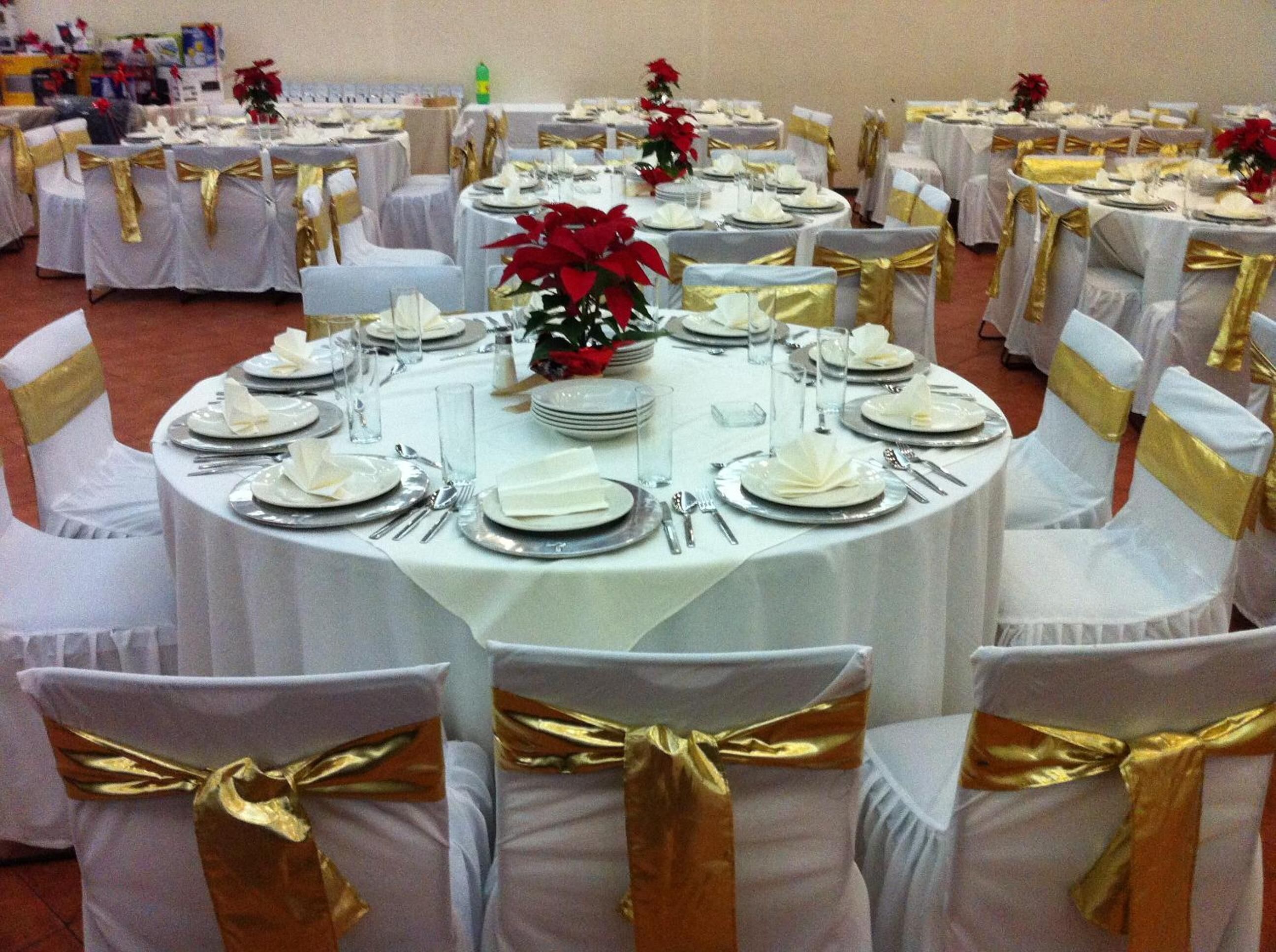 Un salón de banquetes con mesas redondas dispuestas para un evento formal, cada una con manteles blancos y fajas doradas en las sillas.