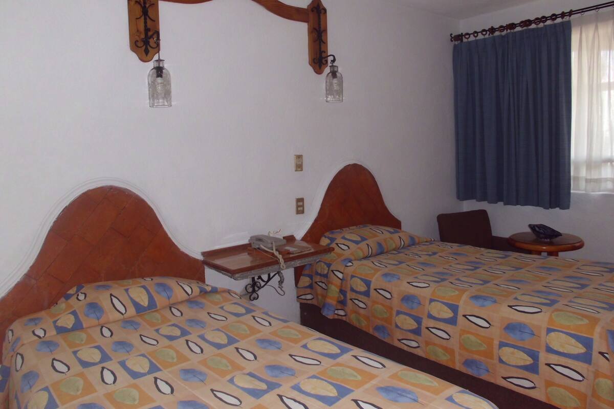 A hotel room with two beds, a small table, a chair, a telephone, and a window with curtains.