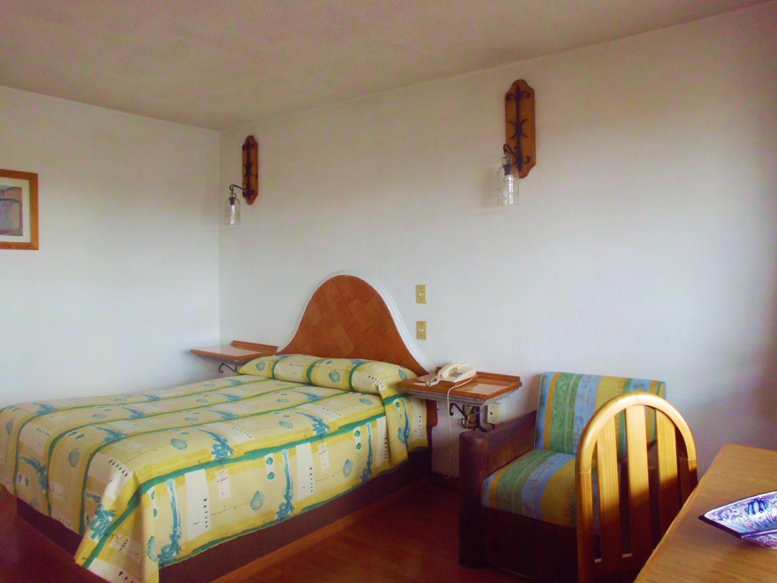 Una habitación pequeña y sencilla con una cama, una silla, una mesa y un teléfono en la pared.