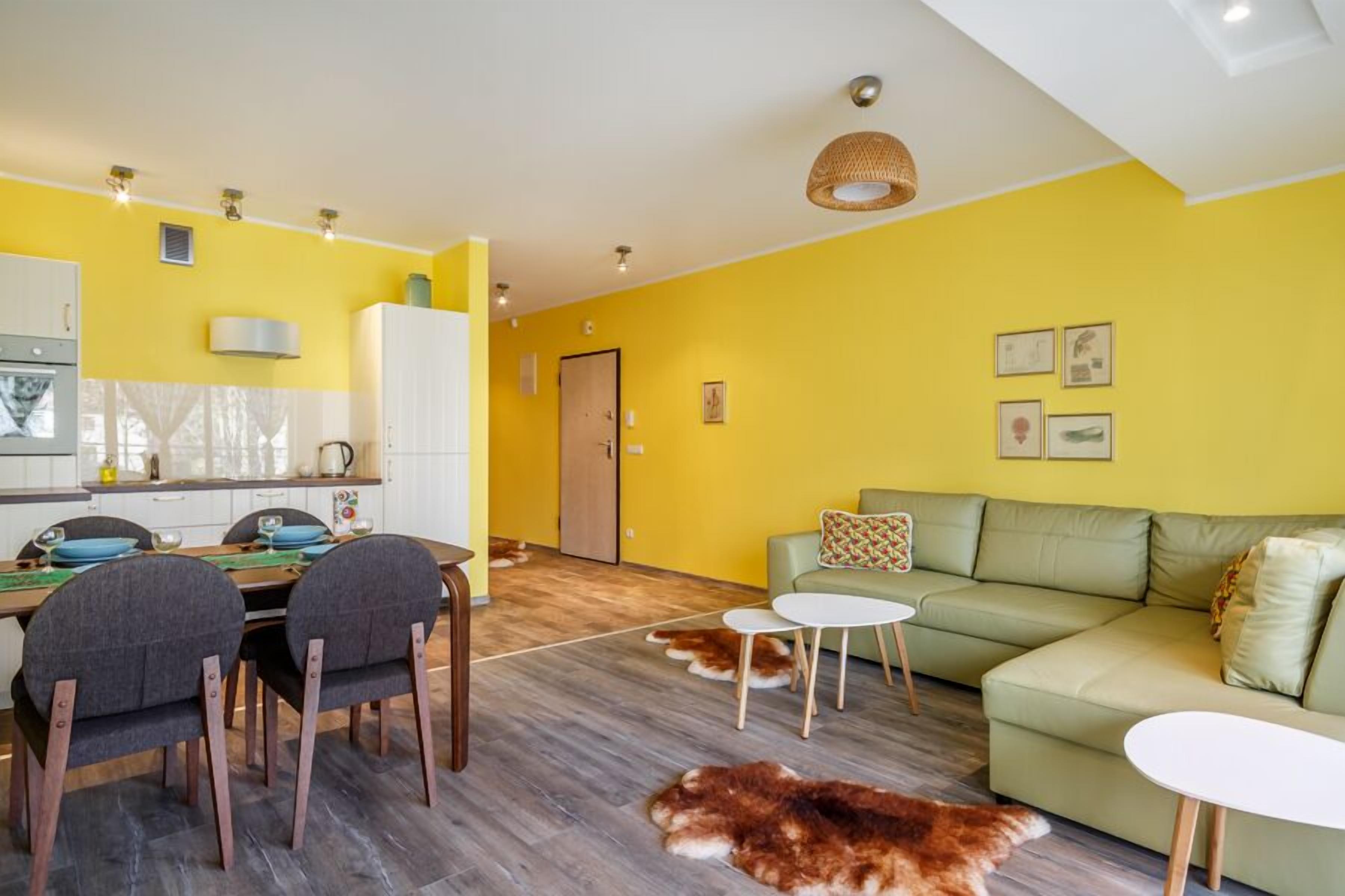 A bright yellow living room with a green sofa, a dining table with chairs, and a kitchen area with white cabinets and a stove.