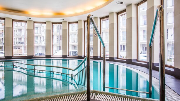Une grande piscine intérieure aux parois en verre transparent, entourée de fenêtres et surmontée d’un plafond incurvé.