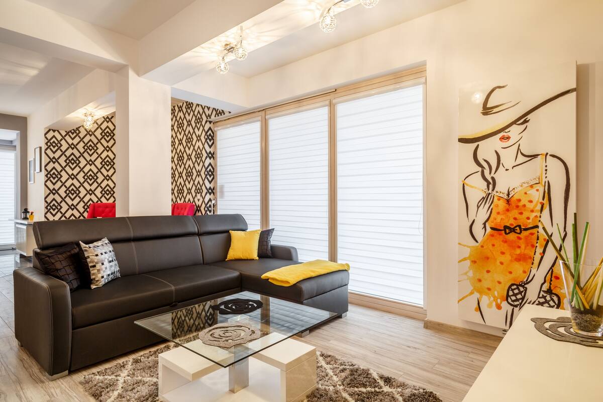Ein modernes Wohnzimmer mit einem schwarzen Ecksofa, einem Glastisch und einem großen Fenster mit Jalousien.