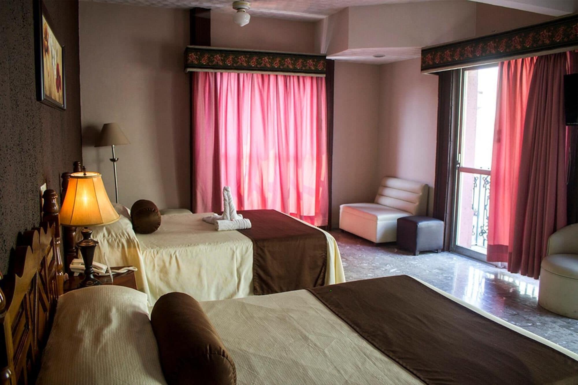 A hotel room with two beds, a sitting area with a chair, a table, and a balcony with curtains.