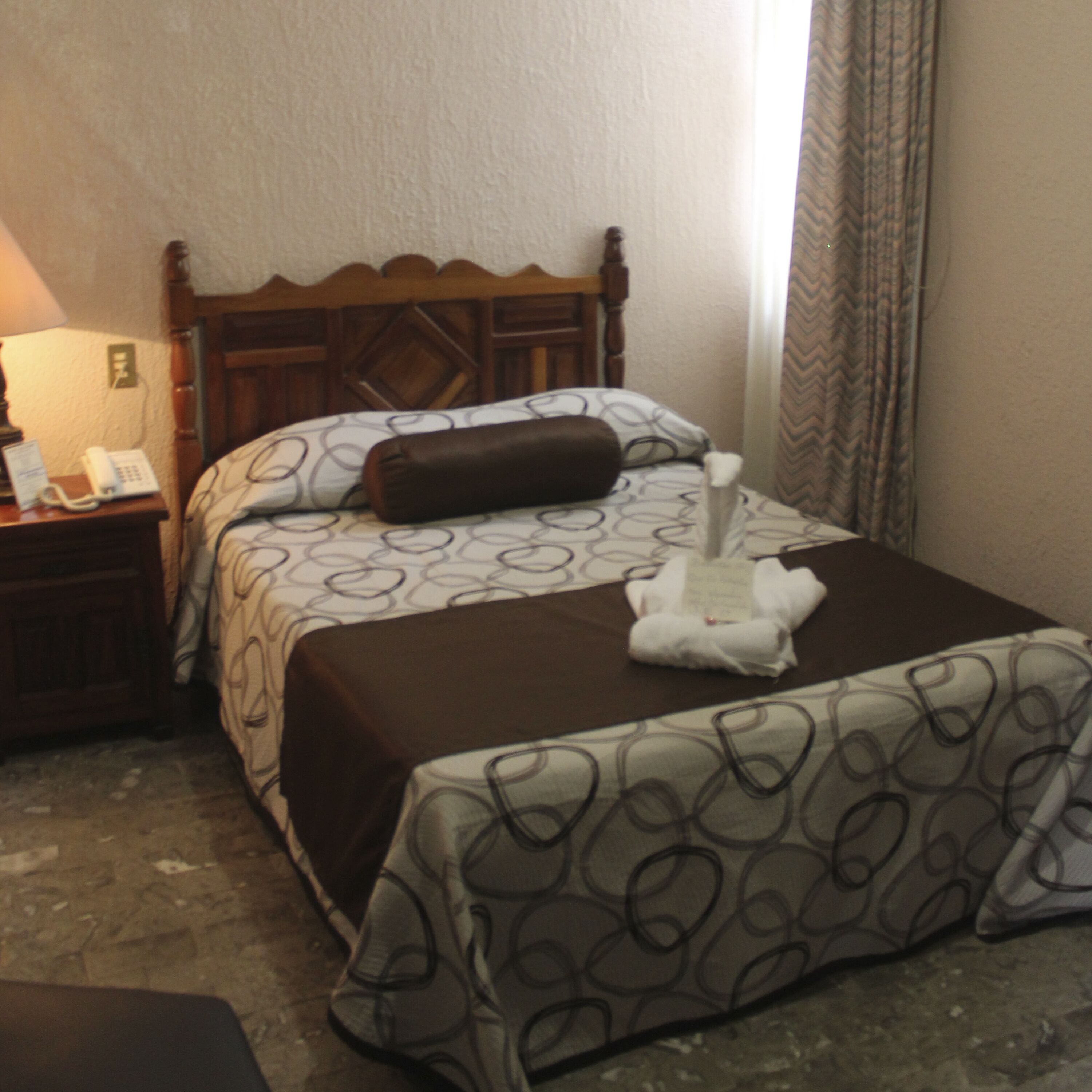 A neatly made bed with a patterned bedspread, a brown pillow, and a folded towel on the bed. There is a wooden nightstand with a lamp and a phone.