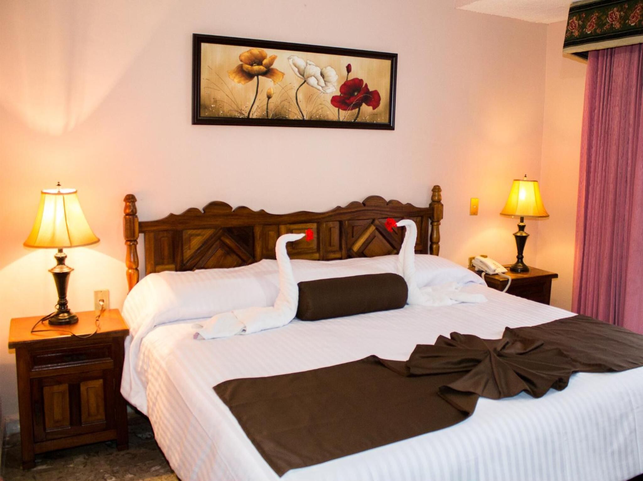 A hotel room with a neatly made bed, two bedside lamps, a wooden headboard, and a floral painting on the wall.