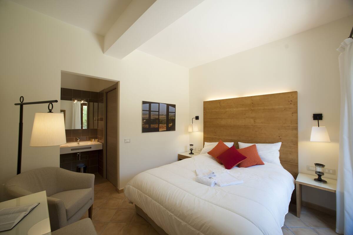 A hotel room with a large bed, a sitting area with a chair, a wooden headboard, a bathroom with a sink and mirror, and a view of the countryside.