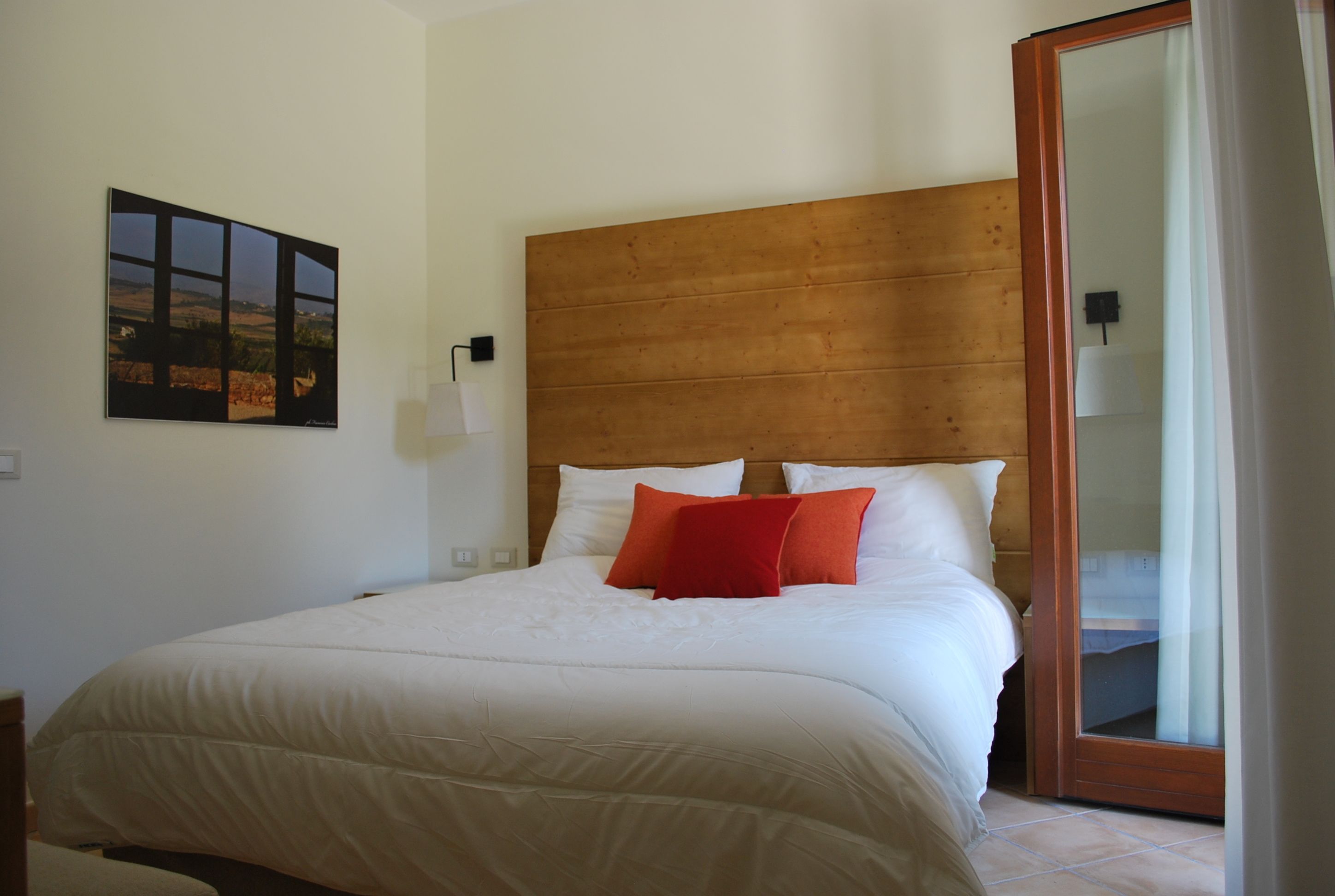 A neatly made bed with white bedding and two red pillows, a wooden headboard, a wall-mounted lamp, and a framed picture.
