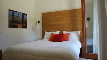 A neatly made bed with white bedding and two red pillows, a wooden headboard, a wall-mounted lamp, and a framed picture.