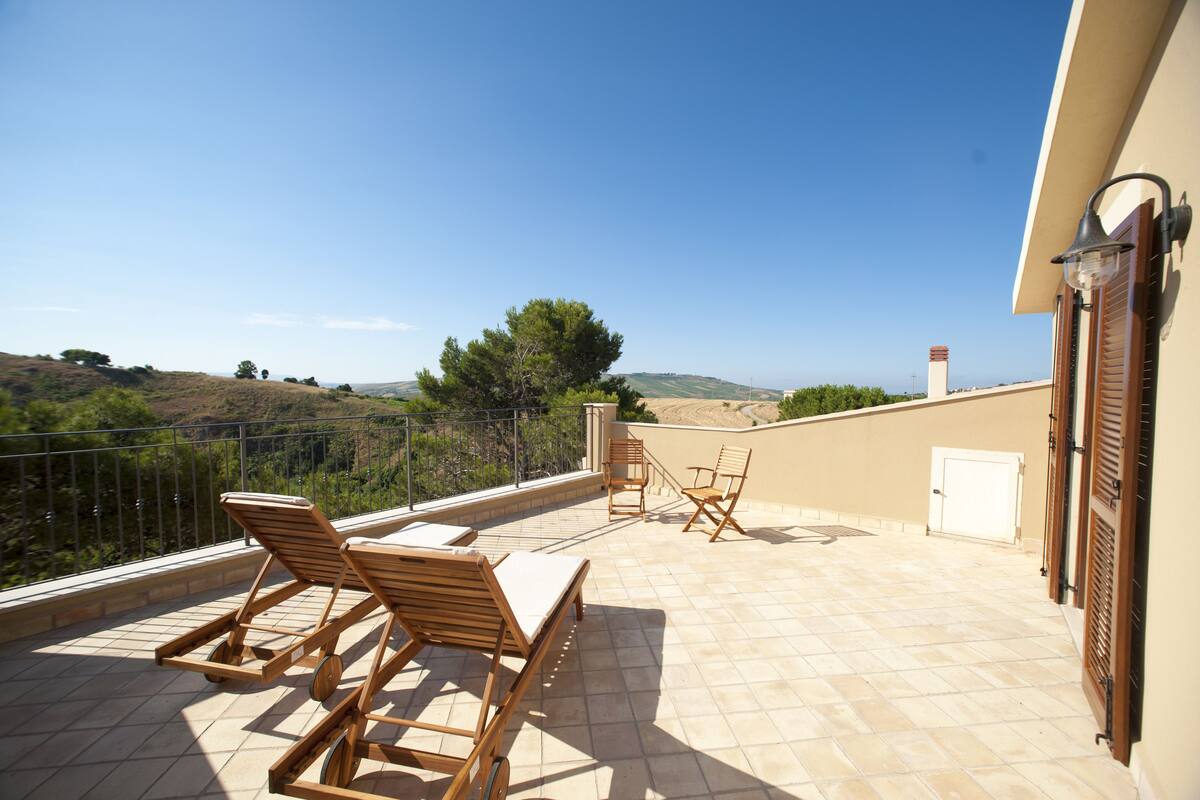 A terrace with wooden lounge chairs and a table, overlooking a hilly landscape.