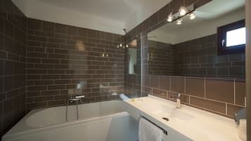 Un bagno moderno con pareti piastrellate di colore scuro, una vasca da bagno bianca, un grande specchio e un lavandino bianco con un asciugamano appeso al lato.