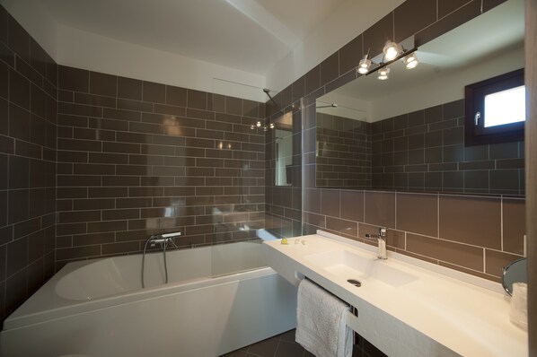A modern bathroom with dark tiled walls, a white bathtub, a large mirror, and a white sink with a towel hanging on the side.