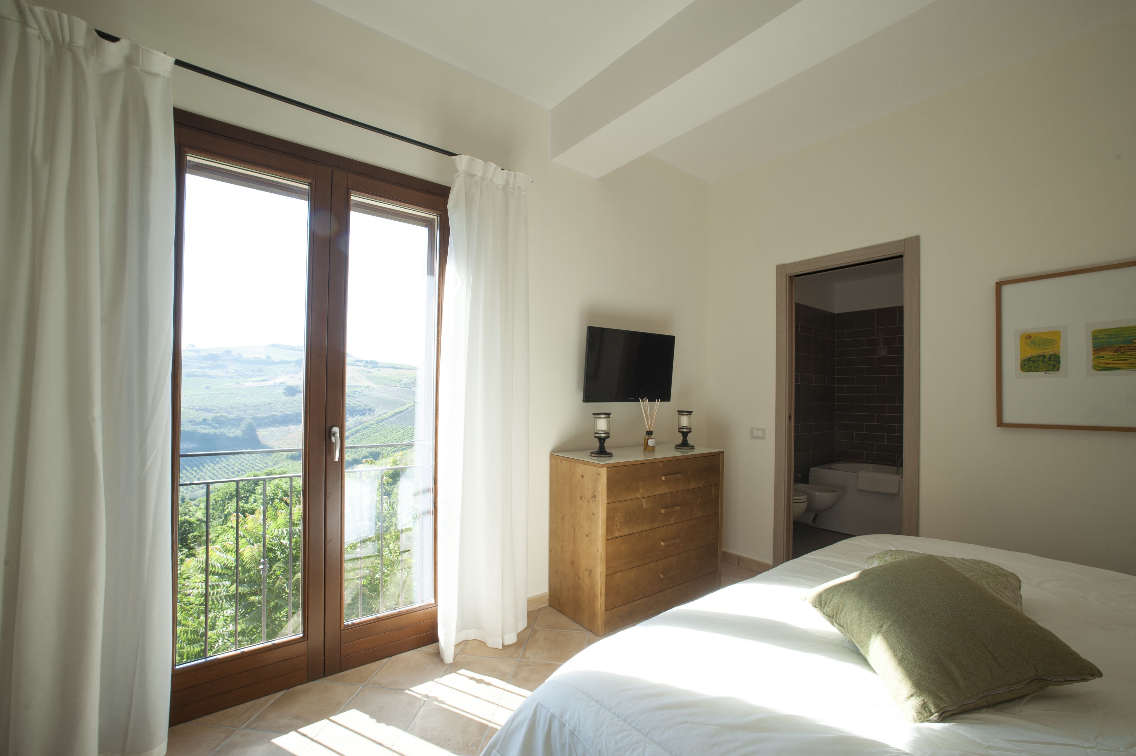 Une chambre à coucher avec un lit, une commode en bois, un téléviseur et une salle de bain visible à travers une porte ouverte.