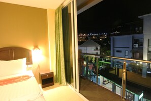 A hotel room with a bed, a nightstand, a window with a view of the city, and a balcony with a railing.