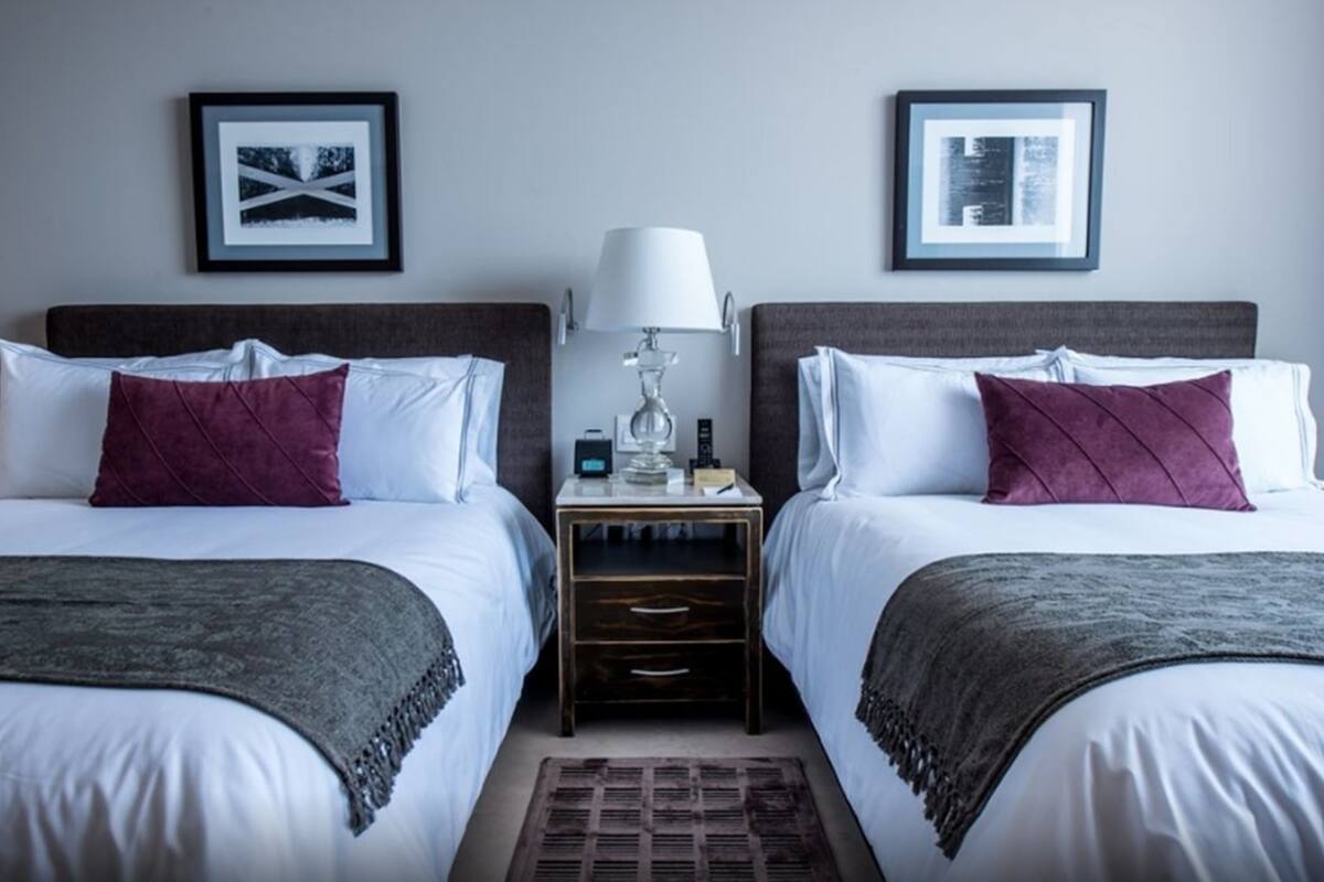 A hotel room with two beds, each with a headboard, a bedside table with a lamp, and a small drawer. There are framed pictures on the wall above the beds.