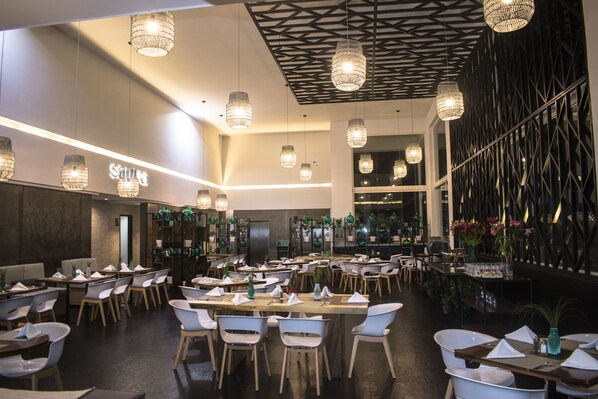 A spacious restaurant with multiple dining tables and chairs, hanging pendant lights, and a decorative wall with a geometric pattern.