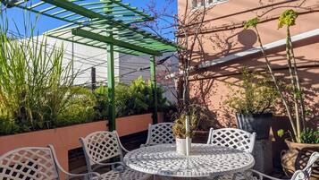 Un patio con sillas y mesas de metal blanco, rodeado de plantas en macetas y una pérgola verde.