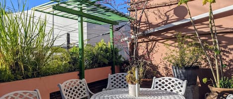 A patio with white metal chairs and tables, surrounded by potted plants and a green pergola.