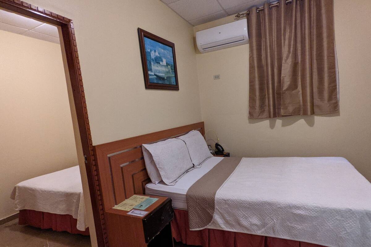 A hotel room with two beds, a wooden headboard, a painting on the wall, an air conditioning unit, and a curtain.
