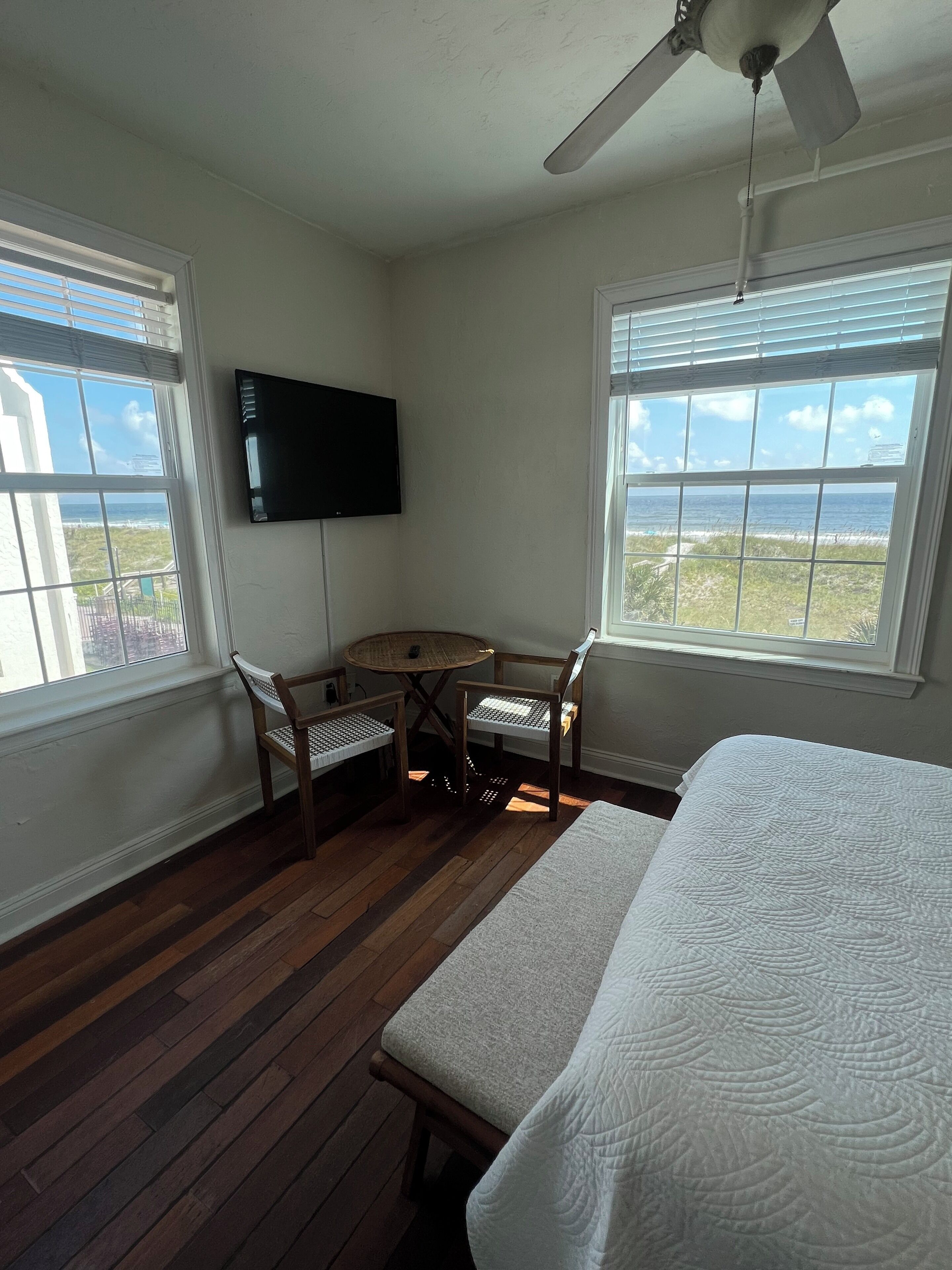 Un dormitorio con cama, ventilador de techo, televisor, una pequeña mesa con sillas y amplias ventanas con vista al océano.