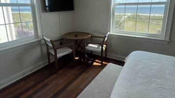 Un dormitorio con cama, ventilador de techo, televisor, una pequeña mesa con sillas y amplias ventanas con vista al océano.