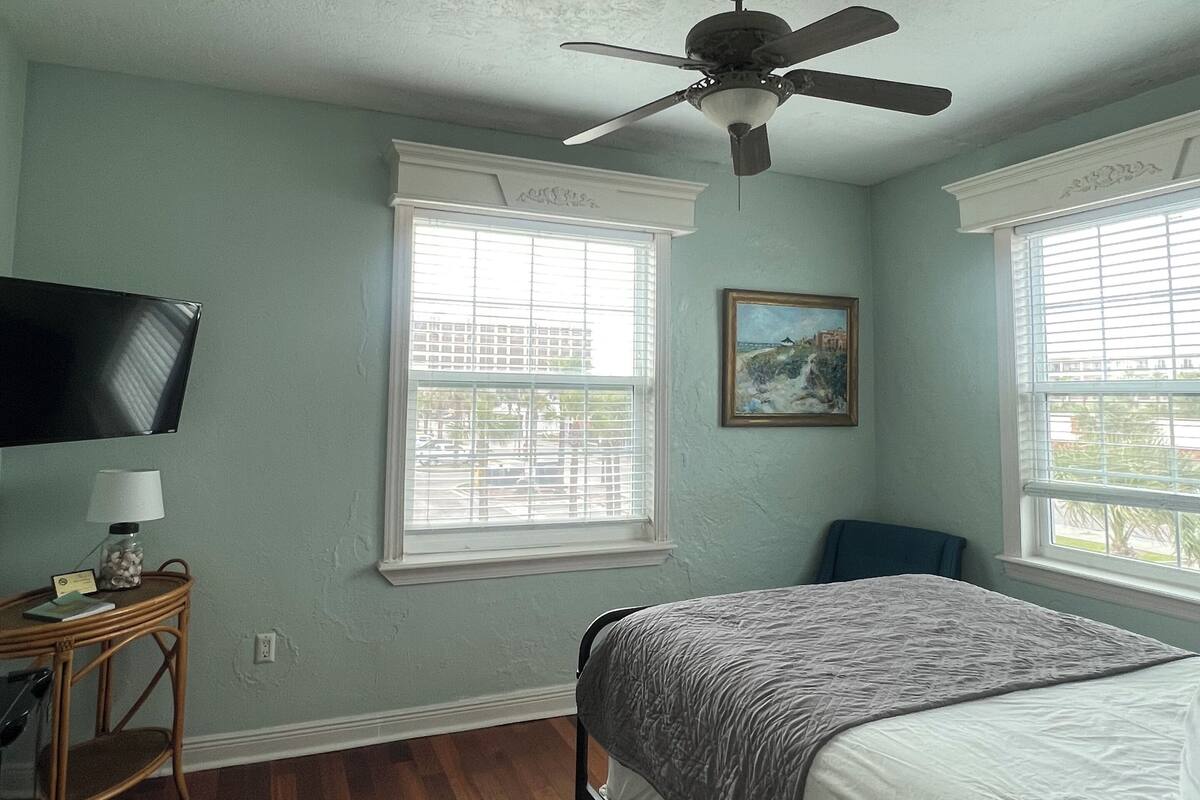 A bedroom with a bed, a ceiling fan, a TV, and a window.