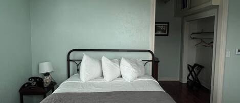 A bedroom with a bed, a nightstand with a lamp, a ceiling fan, and a door leading to another room.
