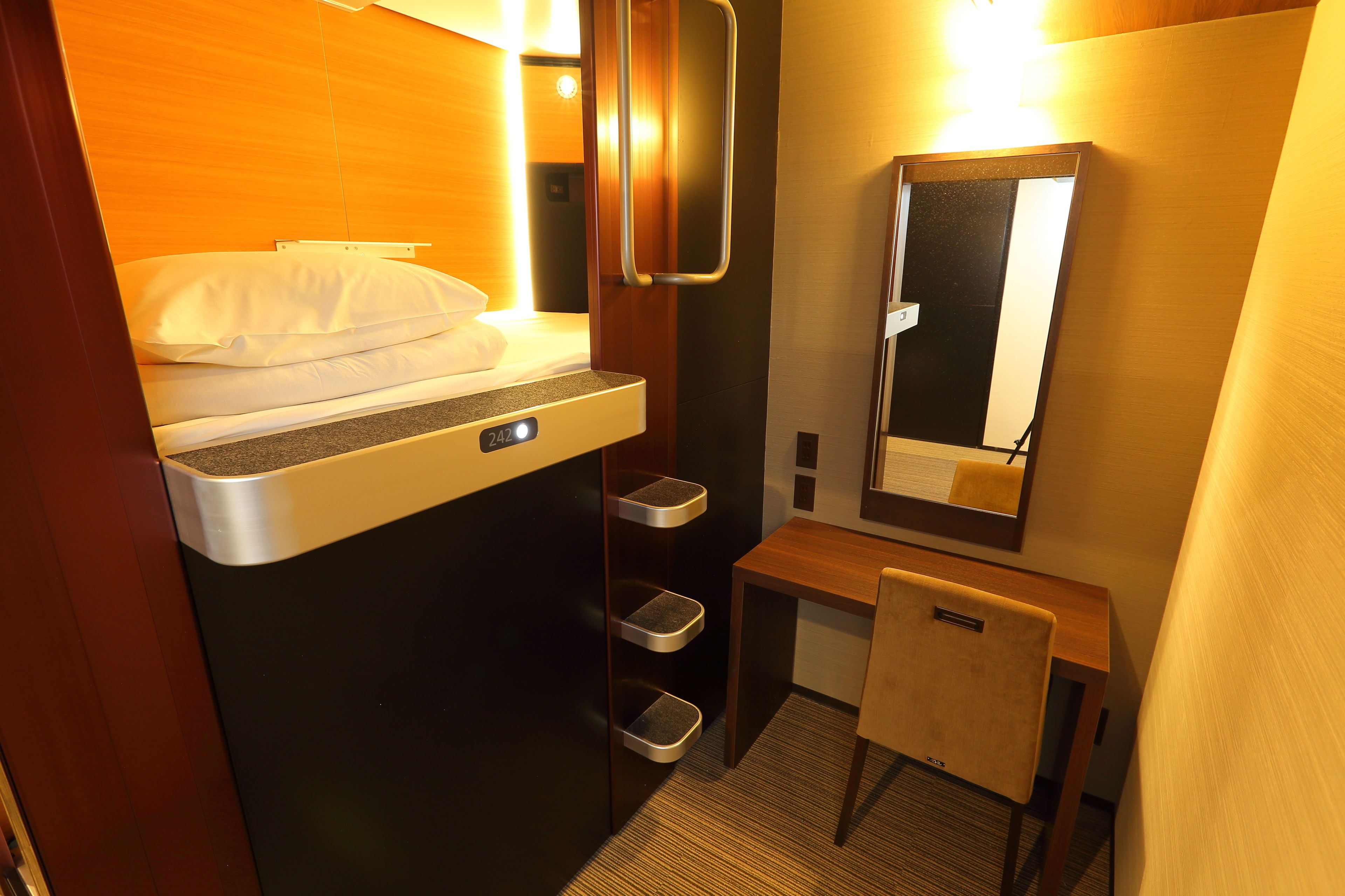 A compact hotel room with a bed, a small desk, a mirror, and a trash bin.