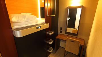 A compact hotel room with a bed, a small desk, a mirror, and a trash bin.