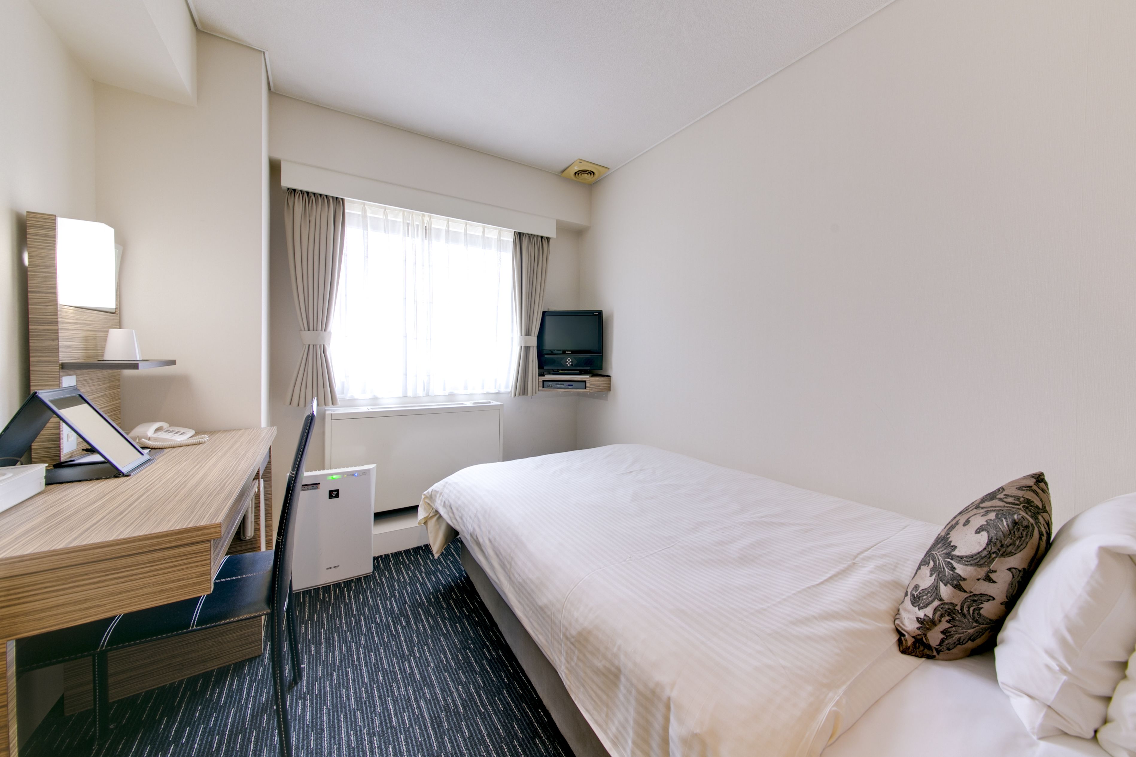 A hotel room with a bed, a desk with a telephone, a television, and a window with curtains.