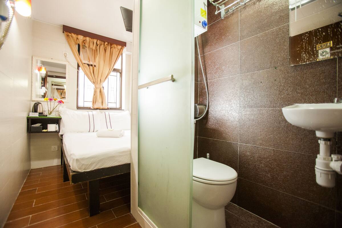 A hotel room with a bed, a bedside table with a lamp, a window with curtains, a bathroom with a toilet and sink.