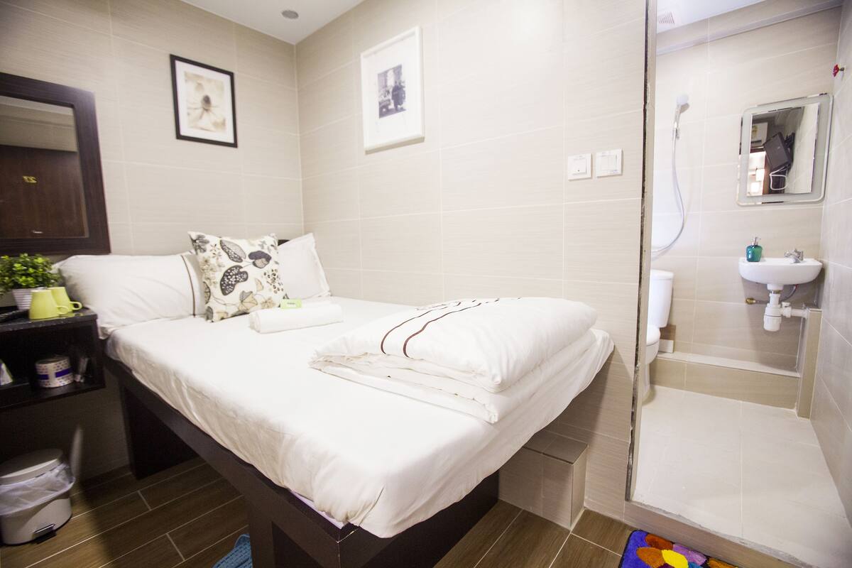 A hotel room with a neatly made bed, a bedside table with a plant, a bathroom with a sink and mirror, and a shower area.