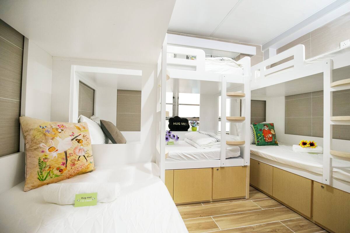 A bunk bed room with a bunk on top and a single bed below. There is a small table with a flower vase and a pillow with a message.