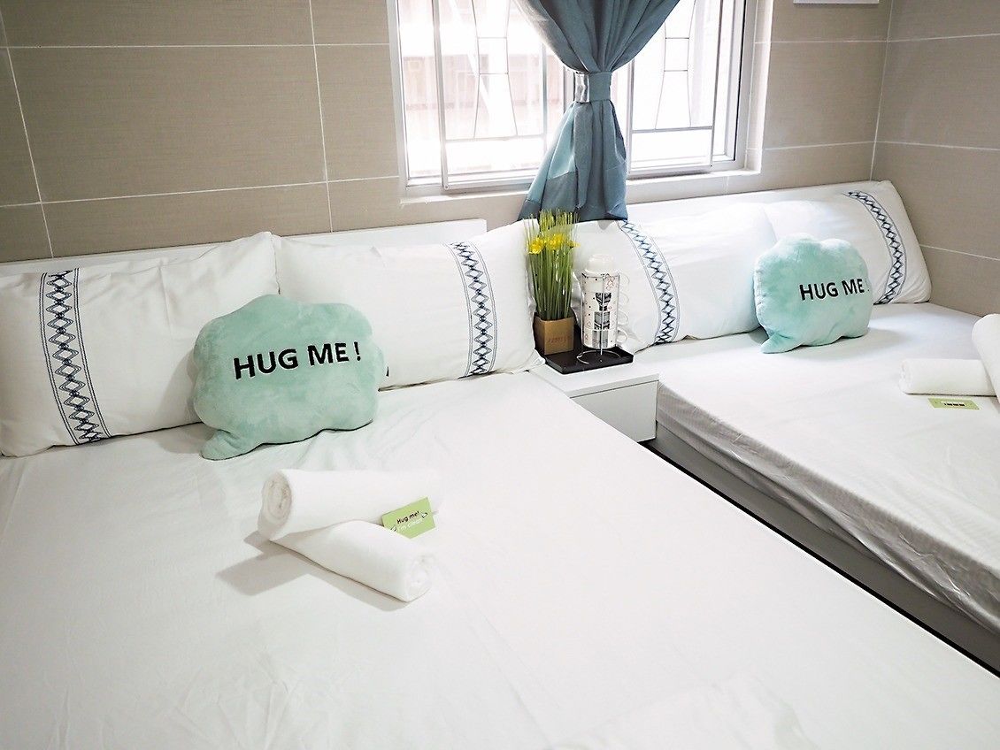 A hotel room with two beds, each with a pillow that says "HUG ME!" and a small table with a vase of flowers.