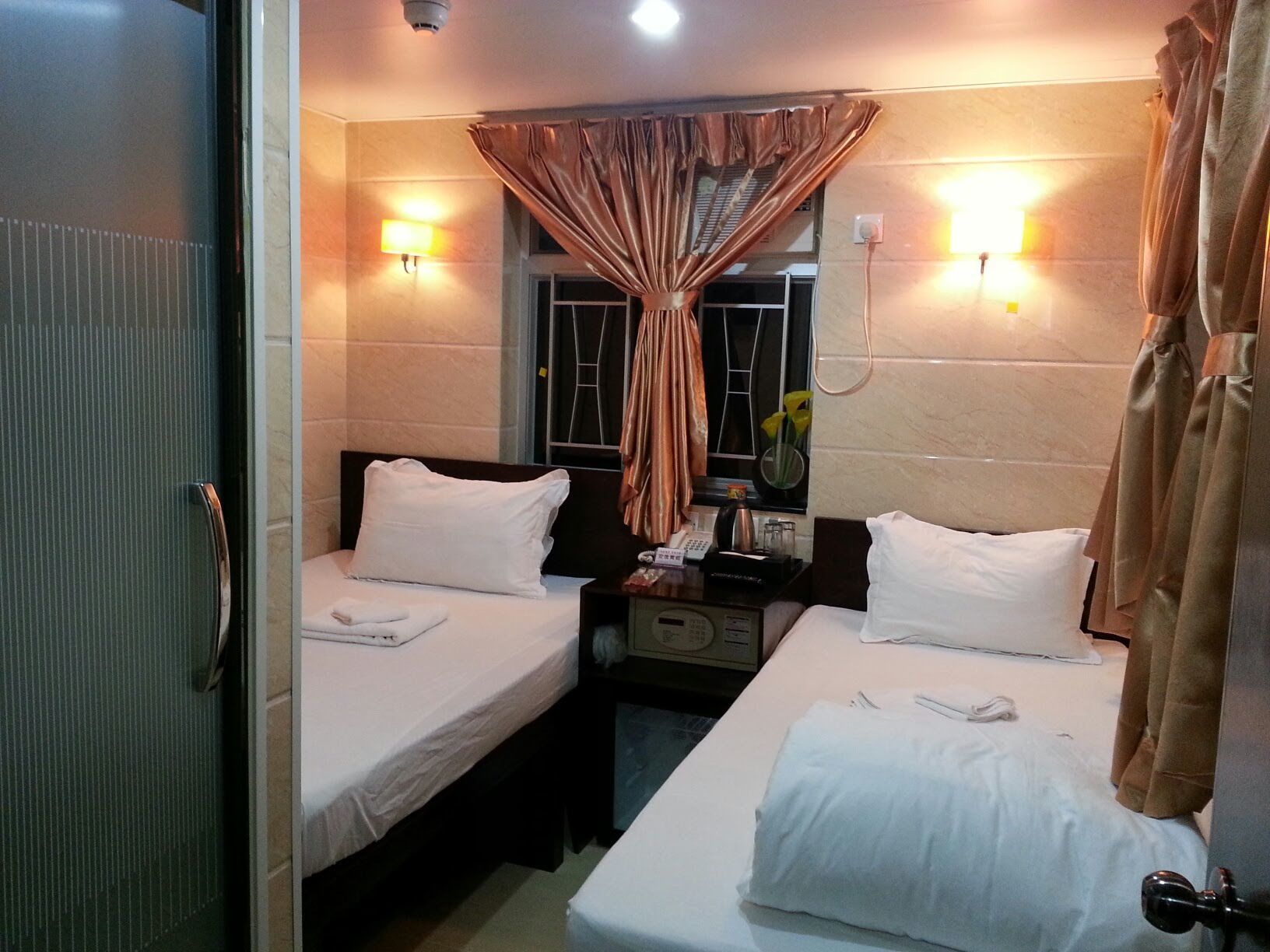 A hotel room with two single beds, a bedside table with a lamp, a window with curtains, and a wall-mounted light fixture.
