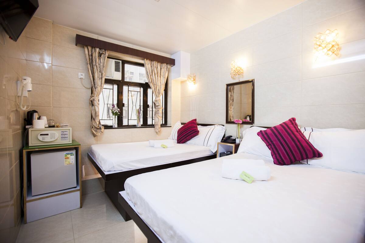 A hotel room with two beds, a small kitchenette, a window with curtains, and a wall-mounted mirror.