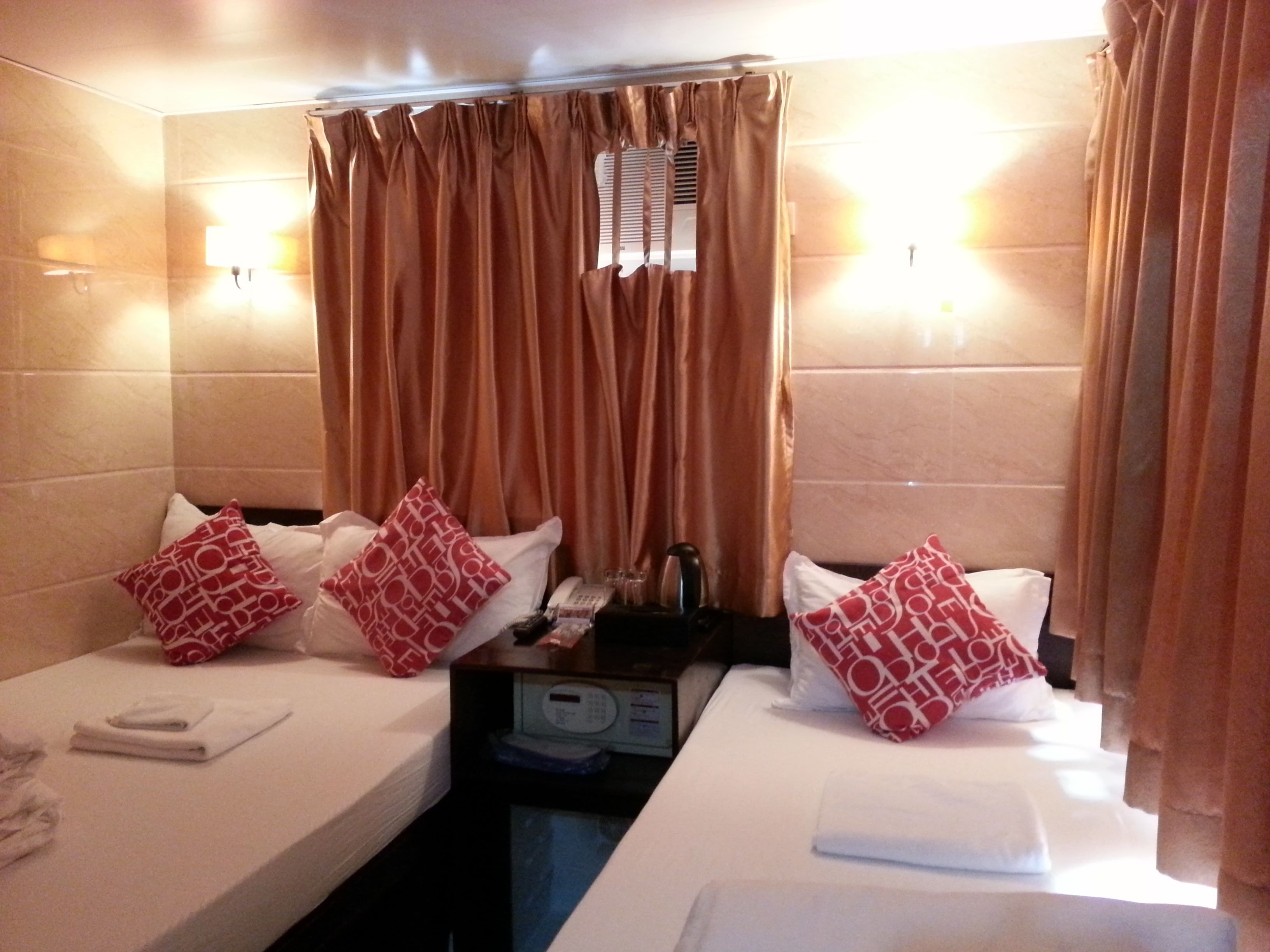A hotel room with two beds, each with a red patterned pillow, a bedside table with a phone, and a small heater.