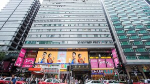 A high-rise building with multiple floors, large windows, and a busy street in front with various shops and taxis. - Comfort Guest House - Hostel (Kowloon)